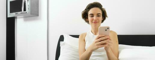 Image of stylish modern woman, sitting on bed, resting at home with laptop and smartphone, sending message, reading notification on mobile phone photo