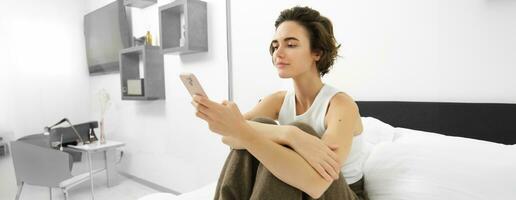 retrato de elegante, moderno joven mujer a hogar, sentado en cama y utilizando móvil teléfono, leyendo en teléfono inteligente, desplazamiento social medios de comunicación solicitud en dormitorio foto