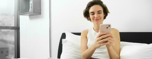 Modern smiling woman in bedroom, sitting on bed, using smartphone and looking at screen, working from home on laptop photo