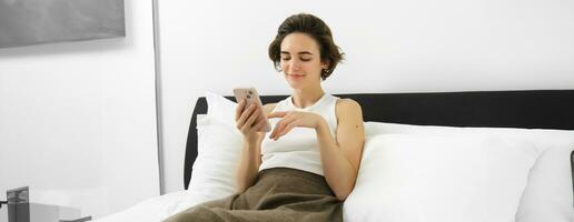 Portrait of cute female model in bedroom, holding phone, typing message on smartphone, resting in bed and smiling photo