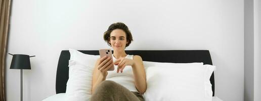 Portrait of young modern woman lying in her bed and using mobile phone, resting in bedroom and looking at smartphone screen photo