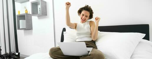 Portrait of happy woman rejoicing, sitting with computer and doing winner dance, has laptop on her laps, raising hands up and celebrating, triumphing with joy photo