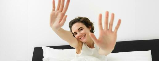 Beautiful young woman stretching her hands towards camera, waking up in morning, resting in her bed, smiling and looking happy photo