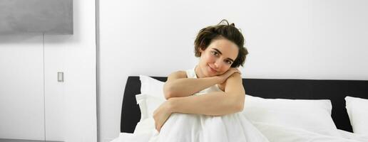 Image of beautiful brunette woman sitting on bed, covered in white linen sheets, looking at camera with dreamy, sensual smile, gazing at someone photo