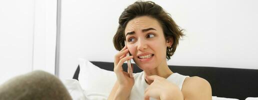 Image of woman with anxious, awkward face expression, talking on mobile phone, cringe from something she hears during conversation, lying in bed, saying something risky photo