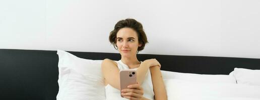 Portrait of smiling brunette woman resting in her bedroom, using mobile phone and lying in bed in her home outfit, messaging, sending message on smartphone app photo