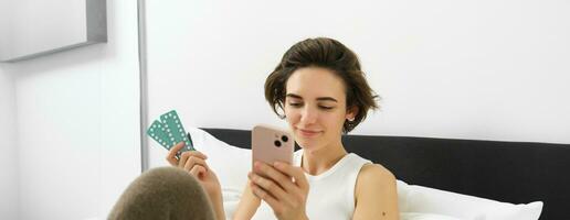 Portrait of smiling young woman reading on mobile phone, searching medication dosage instruction, her prescription on medical app, holding pills, sitting on bed photo