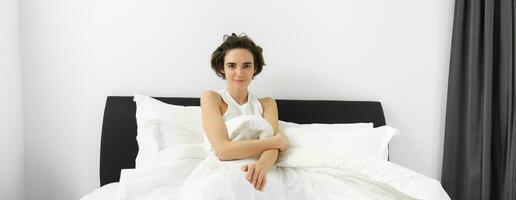 Image of young sensual woman with short hair, wearing tank top pyjama, lying in bed under white linen sheets, looking at camera, waking up in morning photo