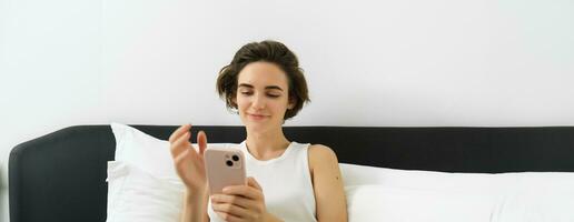 Portrait of young smiling woman resting in her bedroom, lying in bed and using smartphone, online shopping on mobile phone, scrolling social media on device photo