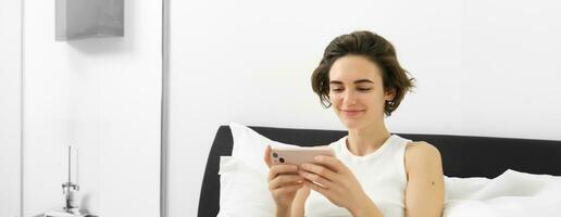 Cute young woman with mobile phone in hands, lying in bed in tank top, looking at smartphone with pleased smile photo