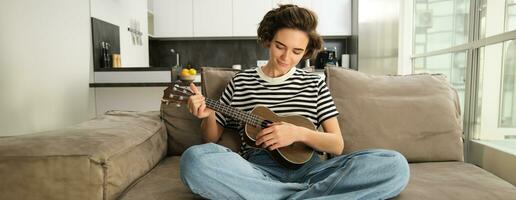 estilo de vida y aficiones concepto. joven sonriente mujer en sofá, jugando ukelele, canto y aprendizaje rasgueo modelo para favorito canción foto