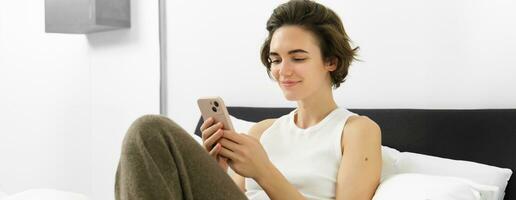 Cute young woman with mobile phone in hands, lying in bed in tank top, looking at smartphone with pleased smile photo