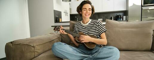 retrato de alegre joven mujer jugando su ukelele, canto y reír, sentado en vivo habitación a hogar foto