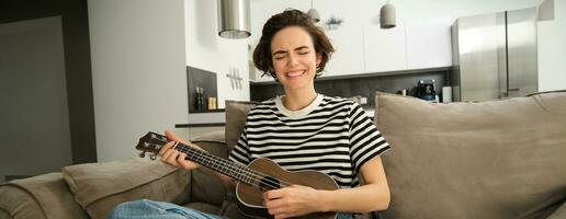 Passionate young woman, musician playing ukulele at home, singing with joy, sitting on couch in living room photo