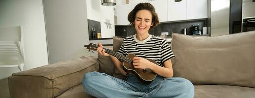 Portrait of cute young woman, girl with ukulele, playing and singing favourite song while sitting in living room on sofa, strumming strings photo