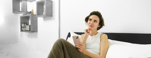 Portrait of young beautiful brunette woman, sitting on bed with smartphone, using mobile phone app, sending message and smiling, reading on telephone application in bedroom photo