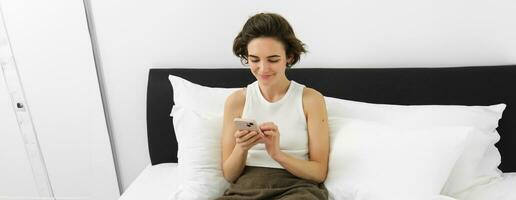Portrait of young beautiful brunette woman, sitting on bed with smartphone, using mobile phone app, sending message and smiling, reading on telephone application in bedroom photo