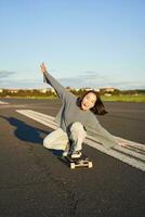 despreocupado patinador niña en su patineta, montando longboard en un vacío camino, participación manos oblicuo y riendo foto