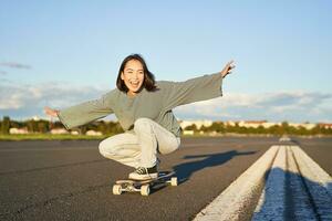 libertad y felicidad. contento asiático niña montando su longboard en un vacío soleado camino, riendo y sonriente, patinar foto