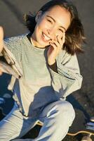 Selfie of beautiful asian girl smiling, taking photo on smartphone while sitting on skateboard outdoors