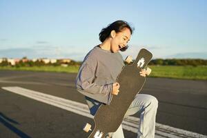 Beautiful asian teen girl playing with her longboard, holding skateboard as if playing guitar, standing on road on sunny day photo