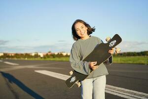 Lifestyle and hobbies. Smiling cute asian girl holding skateboard and walking towards sun on an empty road photo