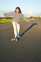 vertical retrato de contento asiático niña disfrutando patineta divertido día afuera. sonriente coreano patinador en longboard, montando a lo largo vacío calle en soleado día foto