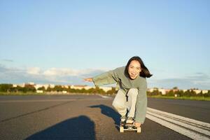 Skater girl riding on skateboard, standing on her longboard and laughing, riding cruiser on an empty street towards the sun photo