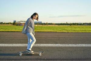 Beautiful asian skater girl riding her longboard on sunny empty road. Young woman enjoying her skate ride smiling and laughing photo