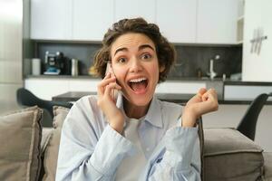 Close up portrait of amazed brunette woman, jumps on sofa from excitement, answers phone call, hear great news over the telephone, reacts surprised and happy photo