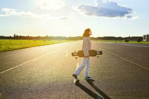 joven patinador chica, adolescente Patinaje en crucero, participación longboard y caminando en hormigón vacío la carretera foto