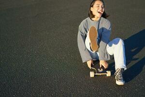 Portrait of beautiful asian girl skating, sitting on her skateboard and smiling. Cute teenager with longboard, skating on road photo