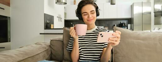 sonriente niña acecho televisión espectáculo en teléfono inteligente, vistiendo auriculares, Bebiendo té y sentado en sofá foto