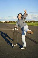 Vertical shot of happy asian skater girl, jumping, standing with skateboard and smiling. Woman skating on longboard and having fun photo