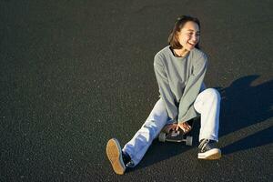 Happy beautiful korean teen girl sits on her skateboard, cruising on longboard, wearing casual clothes photo