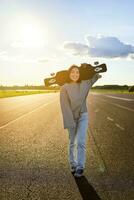 Young asian woman standing with longboard on sunny road, skating in skate park on her cruiser photo