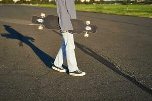 Cropped shot of teen girl body, holding cruiser longboard in hand, walking in sneakers on road in jeans and sweatshirt. Young woman skater with skateboard photo