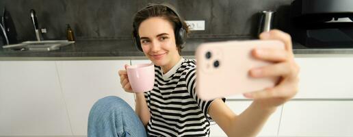 elegante hembra modelo en auriculares, toma selfie con taza de té en el cocina, destino en social medios de comunicación aplicación, utilizando móvil teléfono cámara a hacer contenido para su Blog foto