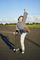 Vertical shot of asian girl feeling excited, skating on longboard, jumping and posing with skateboard, standing with cruiser on empty road, having fun outdoors photo