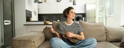 Portrait of girl resting in living room, playing ukulele, learns how to play new musical instrument, spending time relaxing at home photo