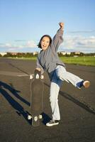 Vertical shot of asian girl feeling excited, skating on longboard, jumping and posing with skateboard, standing with cruiser on empty road, having fun outdoors photo