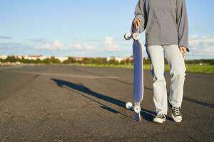 Cropped shot of female body, holding skateboard in hand, walking on road. Young woman carry her longboard, cruiser, skating on the road photo