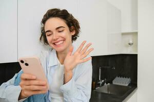 retrato de simpático sonriente morena mujer, sentado a hogar en cocina, mirando a su teléfono inteligente cámara, vídeo chats y olas a móvil teléfono, diciendo Hola a alguien foto