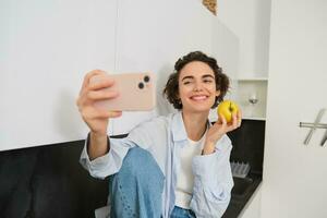 Beautiful girl blogger, takes selfie as she eats apple, shows how to lead healthy lifestyle to followers online, holds smartphone for photo