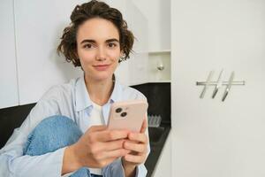 Smiling brunette girl sitting with smartphone at home, orders takeaway from mobile phone app, using online shopping application photo