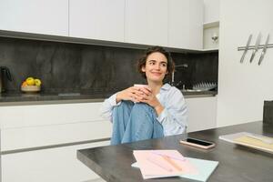 Image of young adult woman, sitting in kitchen with cup of coffee, smiling and looking aside, relaxing at home with cuppa photo