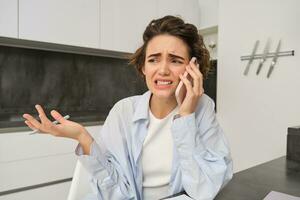 Portrait of woman with guilty face talking on mobile phone, sitting at home and having uncomfortable conversation over the telephone photo