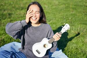 sonriente asiático niña con ukelele, jugando en parque y cantando, estilo de vida concepto foto