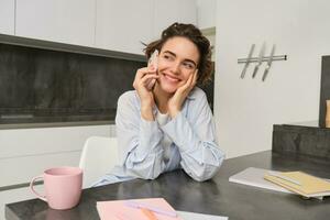 retrato de joven mujer, 25 años viejo, participación teléfono inteligente, hablando en móvil teléfono, sentado en cocina a hogar foto