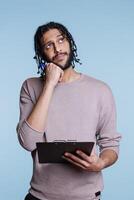 Pensive arab man thinking and looking away with dreaming facial expression while holding clipboard. Thoughtful young person planning and writing checklist while standing in puzzled pose photo
